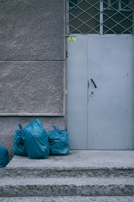 The image shows four blue rubbish bags placed on a concrete step in front of a grey metal door with a black door handle, set into a building with a textured, grey concrete wall. To the right of the bags, the door features a small lock and a handle, with a diamond-patterned metal grille covering a window above it, suggesting a secure entrance or service area. The bags appear to be made of plastic and are tightly tied at the top, indicating they are ready for collection or disposal. The concrete surface beneath the bags is slightly weathered, and the overall environment appears clean but utilitarian, typical of an exterior access point for waste collection. This scene on the building's exterior is relevant to private rubbish removal services, where independent collection of rubbish bags from designated access points supports alternative waste handling outside of local authority methods. Rubbish Removal Barnes may assist with clearing similar waste accumulations efficiently, offering an organized approach to rubbish disposal in urban settings.