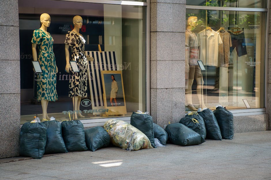 A row of dark green refuse bags and one larger transparent bag filled with mixed waste are lined up on a paved sidewalk outside a clothing shop, which has large display windows showcasing mannequins dressed in patterned dresses and casual shirts. The bags are tightly tied at the top with yellow and white drawstrings, and are positioned directly in front of the shop's glass windows. Inside the window, mannequins are dressed in vibrant and neutral-toned clothing, with some items on display racks. The shop's exterior features a stone framing around the windows, and the scene is lit naturally, with reflections visible on the glass. The placement of the bags suggests an outdoor rubbish collection, possibly an example of off-site waste removal carried out by a private service like Rubbish Removal Barnes, serving businesses on Barnes High Street.