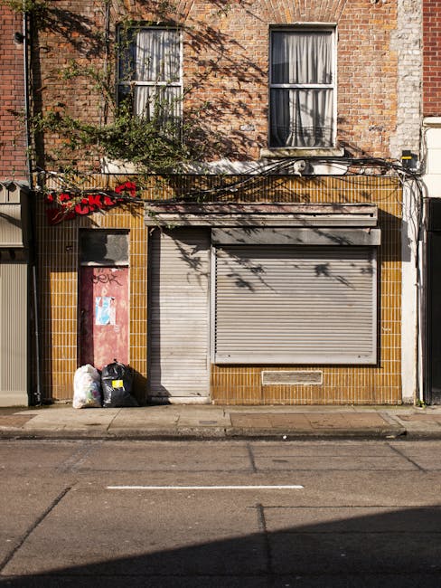 A close-up view of the ground level of a brick building facade, showing a closed metal roller shutter door painted in a light grey tone, with a small vent beneath it. To the left of the shutter, there are two black plastic rubbish bags and a white bag with yellow markings placed on the pavement, indicating waste collection. The building’s exterior features a mix of deep yellow tiles around the door area and exposed reddish-brown brickwork above, with weathered patches and some ivy or plant growth adjoining the left side. Two large windows with black frames and cream-colored curtains are visible on the upper floor, casting shadows onto the brickwork due to sunlight. Overhead, a network of black cables and wiring runs horizontally across the storefront, and the overall scene suggests a typical commercial or retail property on an urban street. This image is relevant for illustrating private waste disposal and rubbish removal services for businesses, as provided by Rubbish Removal Barnes, focusing on the need for on-site clearance of waste accumulated outside shop fronts.