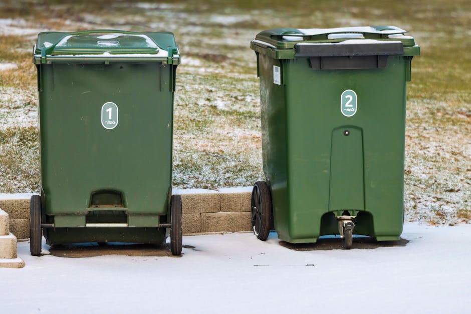 Two green wheeled rubbish bins are positioned side by side outdoors on a concrete surface with a low brick border in the background. The bin on the left has a slightly rounded lid with white markings and a small, black pedal at the base for opening. The bin on the right features a flat, dark-colored lid secured with black hinges, and a black wheel at the back for easy mobility. Both bins have white circular labels with black numbers, with the left marked '1' and the right marked '2,' indicating different waste collection categories. The environment appears to be a driveway or similar outside area, with sparse grass or ground visible beyond the brick border. Light snow covers parts of the surface beneath and around the bins, suggesting a cold season. The scene is evenly lit, highlighting the smooth, plastic surface of the bins and their durable finishes, supporting the context of private waste management or alternative rubbish collection methods often handled by local waste removal services such as Rubbish Removal Barnes.