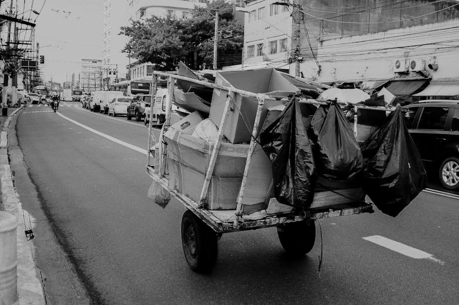 A small, wheeled cart loaded with a variety of discarded items is positioned on a city street in this black and white image. The cart is constructed with a metal frame and has two large, solid rubber wheels at the rear, supporting a flat platform. On the platform, there are several large cardboard boxes, some appearing torn or crushed, filled with miscellaneous debris and household waste. Attached to the sides of the cart are multiple black plastic garbage bags, tied at the top and hanging loosely, likely containing additional waste. The scene is set on the edge of a busy urban road, with parked cars lining the curb on the right side and moving vehicles seen in the distance. Large apartment buildings and commercial structures are visible in the background, with overhead utility wires stretching across the scene. The overall environment suggests an area where private waste collection services, such as those provided by Rubbish Removal Barnes, might undertake on-site clearance of accumulated rubbish, highlighting the importance of independent collection methods for waste management in urban settings.