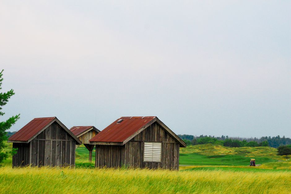 A rural landscape featuring three small wooden sheds with weathered, dark gray planks and rusted reddish-brown metal roofs, positioned amidst a lush, green grassy field. The structures are closely grouped in the foreground, with one shed having a small, white louvered window on its side, and the others showing simple, sloped rooflines without additional visible features. To the left, partially visible, is a green coniferous tree. The background displays a wide expanse of rolling green fields extending into the distance, bordered by a line of trees and shrubs under a pale, overcast sky. On the far right, a small tractor is seen working on the grass, adding a subtle element of active farm or grounds maintenance. The scene communicates a peaceful rural setting potentially associated with independent waste handling or on-site clearance activities, consistent with private rubbish removal services such as those provided by Rubbish Removal Barnes.