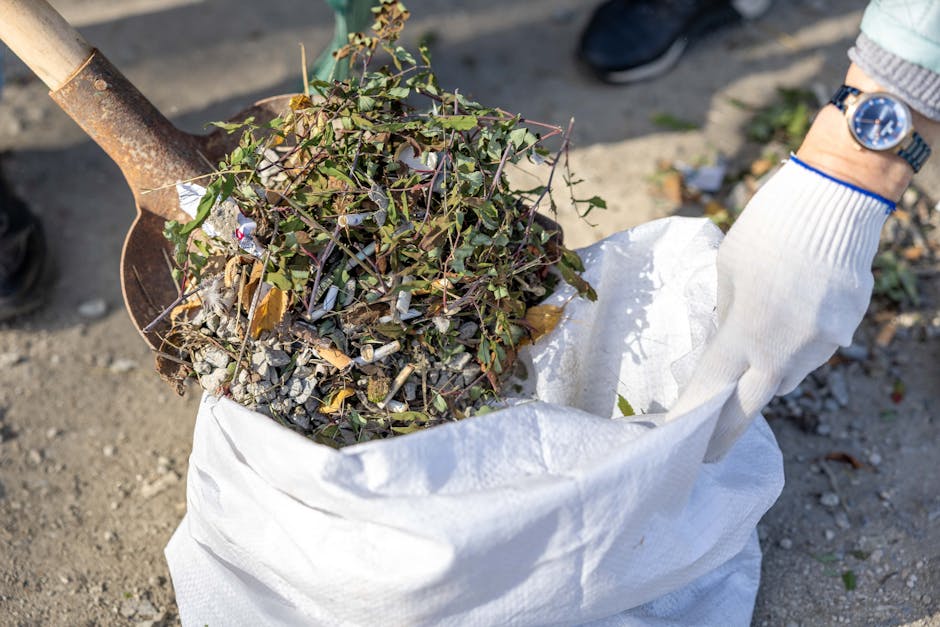 A person wearing a white glove with a blue wristwatch is holding an open white refuse sack filled with a mixture of garden waste, including small branches, leaves, and twigs with green and brown foliage. The sack appears to be made of a durable woven material and is resting on a gravel surface outdoors. Nearby, a rusty metal spade with a wooden handle is partially embedded in the ground, suggesting ongoing gardening or clearance work. In the background, there is another person wearing dark shoes and standing on a paved or gravel area, indicating a garden or outdoor space suitable for waste collection or disposal. The natural light casts shadows on the ground, highlighting the textures of the garden debris and the packaging. The scene exemplifies a typical task involved in waste clearance services, such as those offered by Rubbish Removal Barnes, which handle collection of garden and household waste for private disposal, aiding in environmentally responsible rubbish removal for SW13 homes near Barnes Bridge Station.